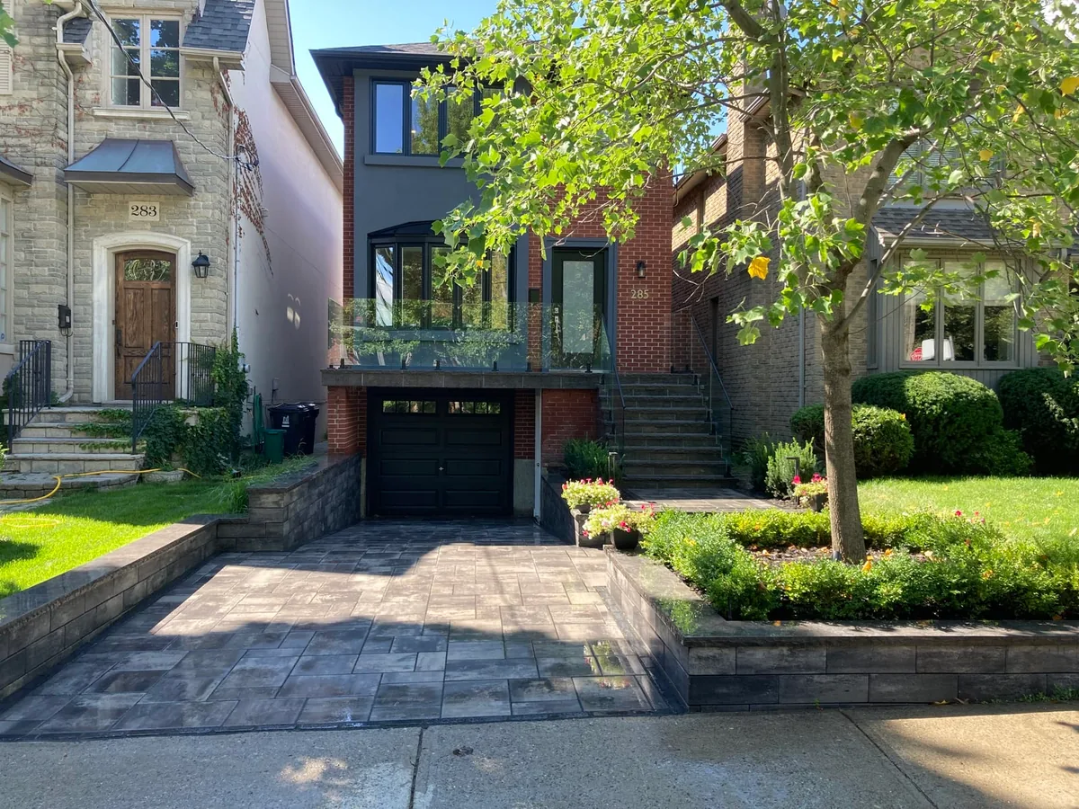 A paver driveway and stone front steps with a glass railing, bordered by stone garden walls and boxwood garden beds in front of a brick home.