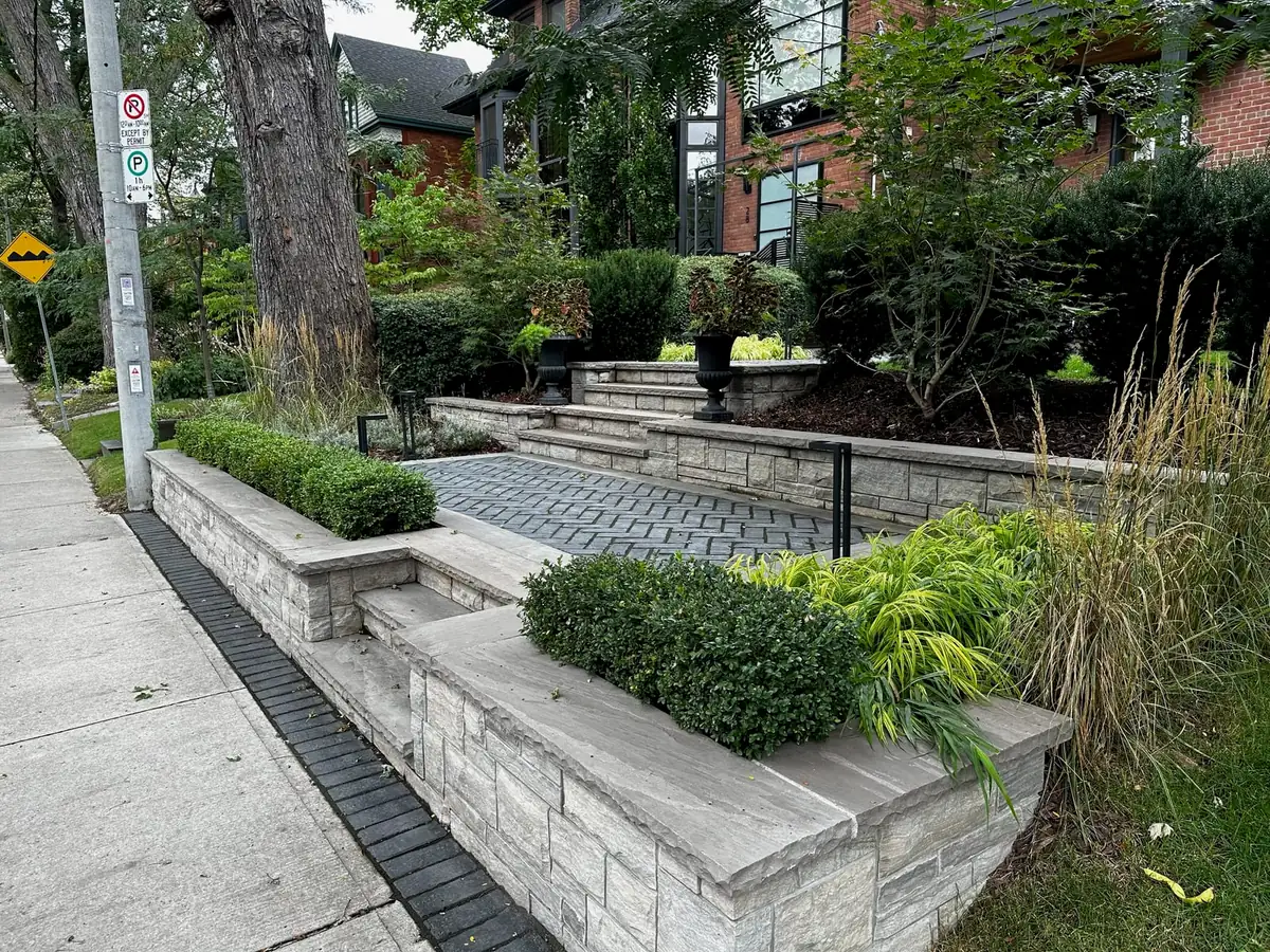 A stone retaining wall bordering a garden bed next to a herringbone-pattern paved driveway with steps.