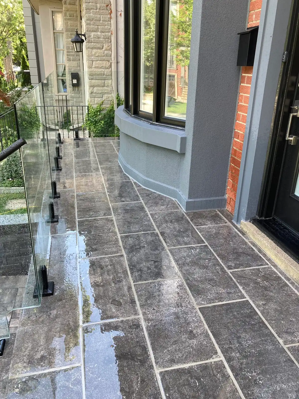 A stone patio walkway along the side of the house with a glass railing and a bay window base.